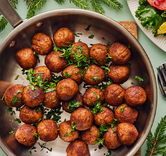 Cooked meat or vegetable meatballs in a stainless steel pan, sprinkled with fresh chopped parsley, on a green background with a side salad dish.