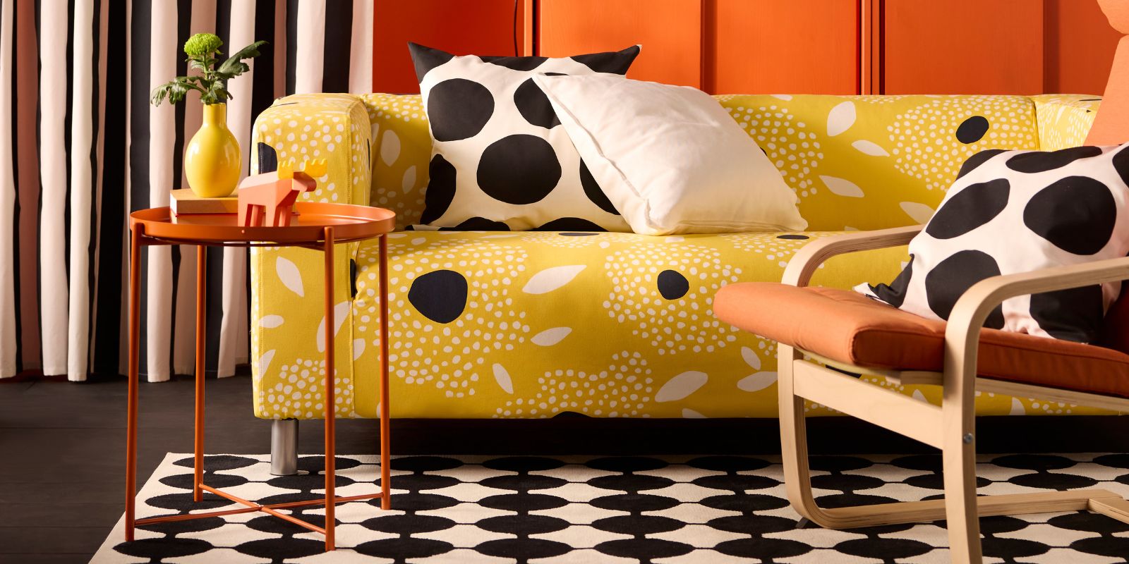 Yellow sofa with black/white patterned pillows. Orange side table with yellow vase. Black/white patterned rug. Chair with orange cushion.