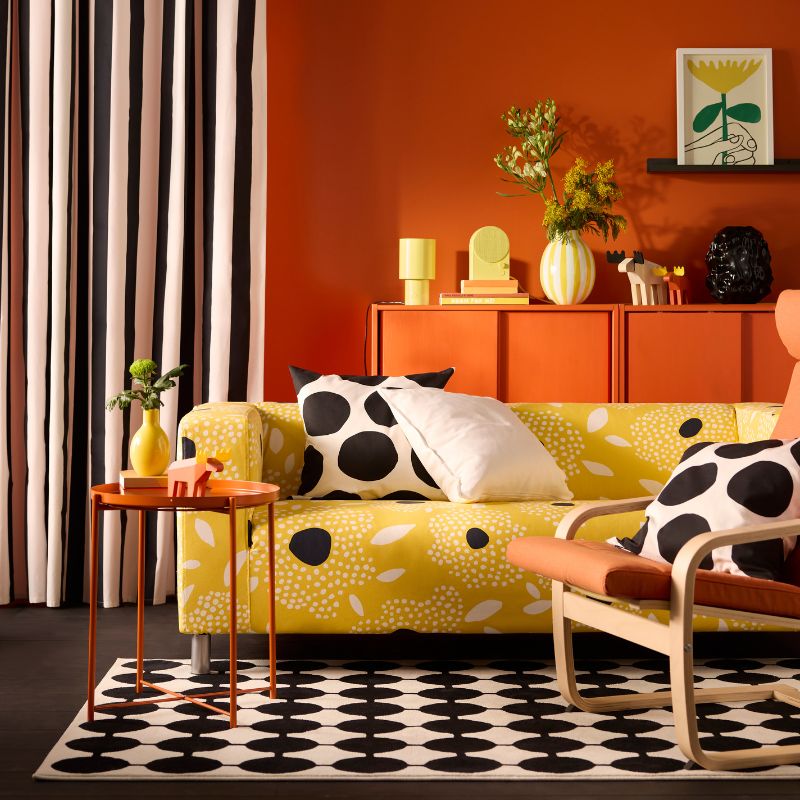 Bright yellow sofa with patterned pillows, orange side table with yellow vase, striped curtains, and a black/white patterned rug.