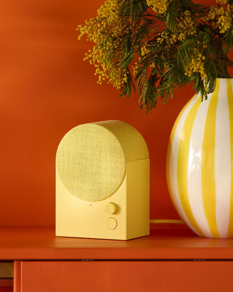 A yellow speaker with a round speaker cover sits next to a yellow and white striped vase holding yellow mimosa flowers.