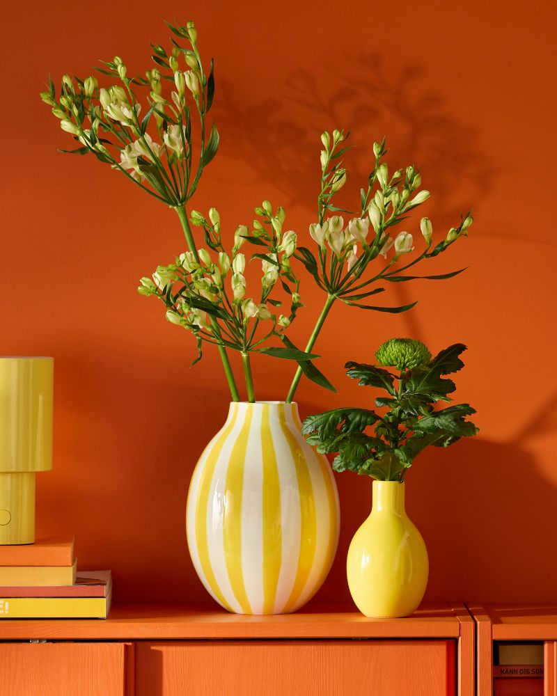 Two yellow vases, one striped and one solid, hold white flowers and green plants on an orange cabinet against an orange wall.