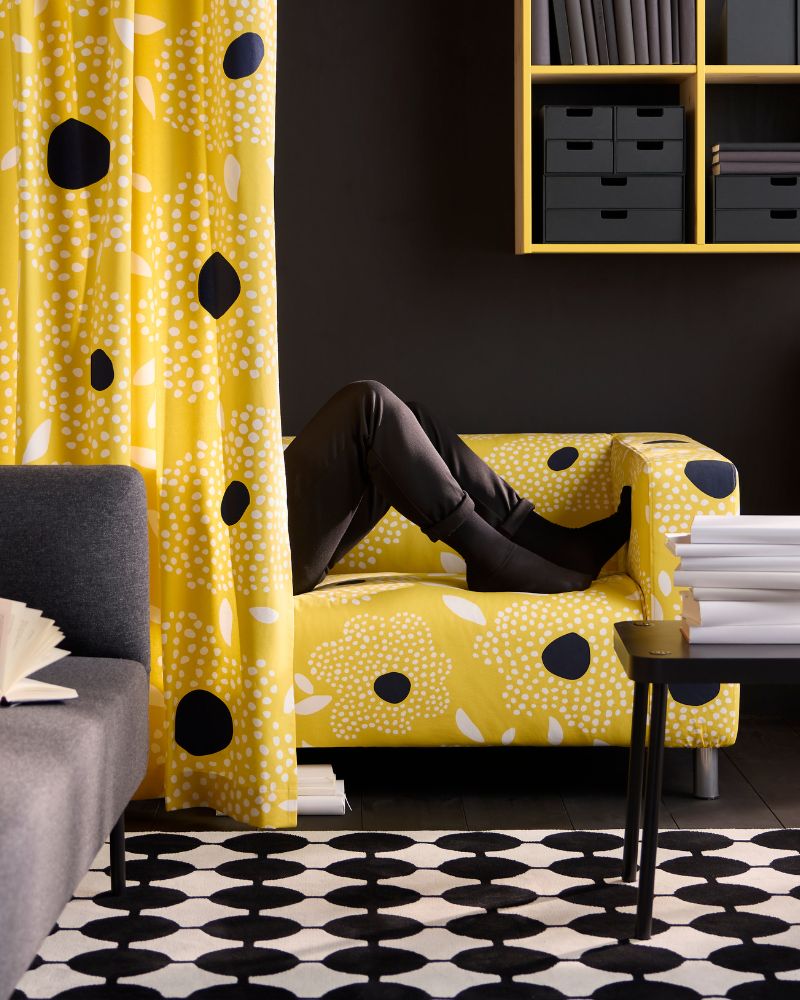 A person's legs are visible on a yellow floral sofa. A grey sofa, black and white rug, and yellow shelves with black boxes are visible.