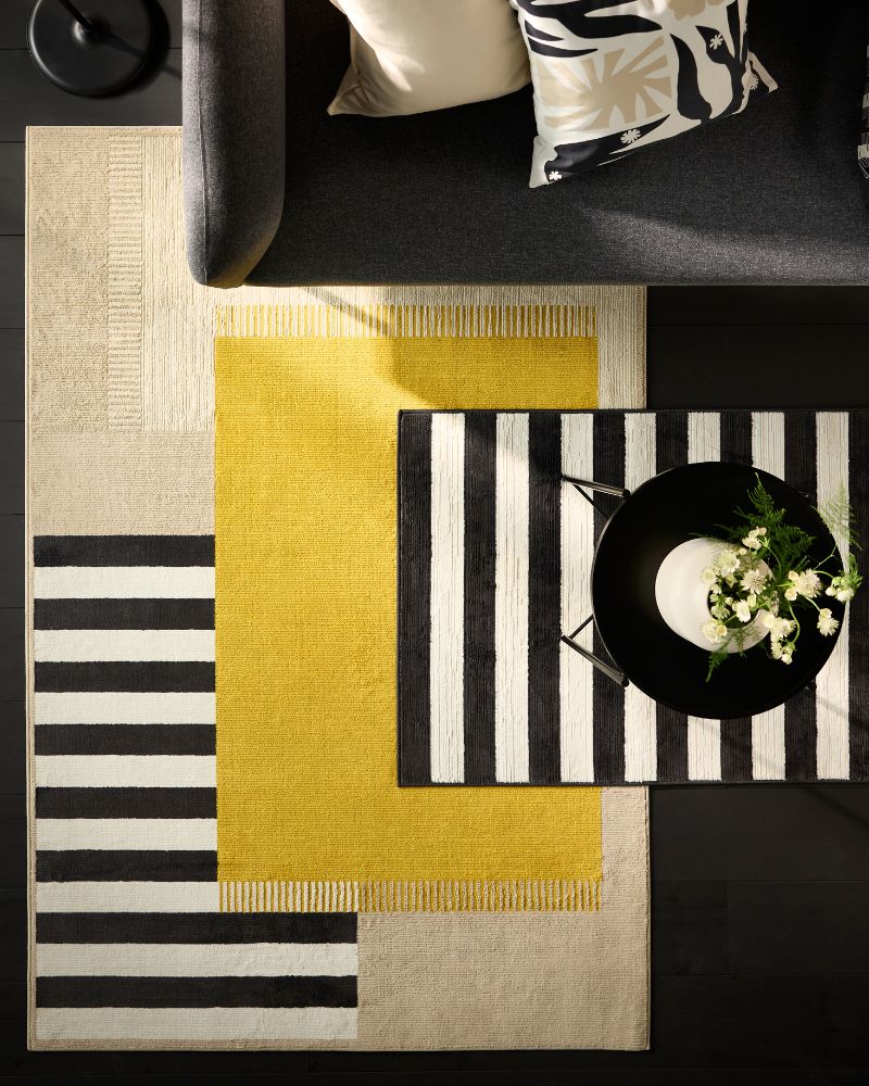 Overhead view of a black sofa with patterned pillows. Three layered rugs (beige, yellow, black/white stripes) under a black coffee table.