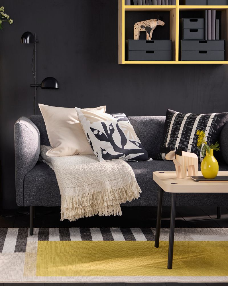 A grey sofa with neutral pillows and a fringe throw. A black floor lamp, a yellow table, and yellow shelves with black boxes are visible.