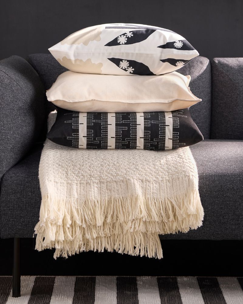 A close-up of a grey sofa with a light beige throw blanket with fringe and a stack of three pillows in neutral and black/white patterns.