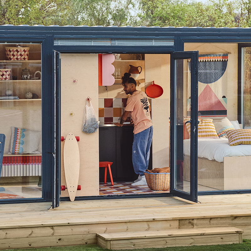 Small house made from shipping containers, with windows to the bedroom and living room; open door shows IKEA OMMJÄNGE items.
