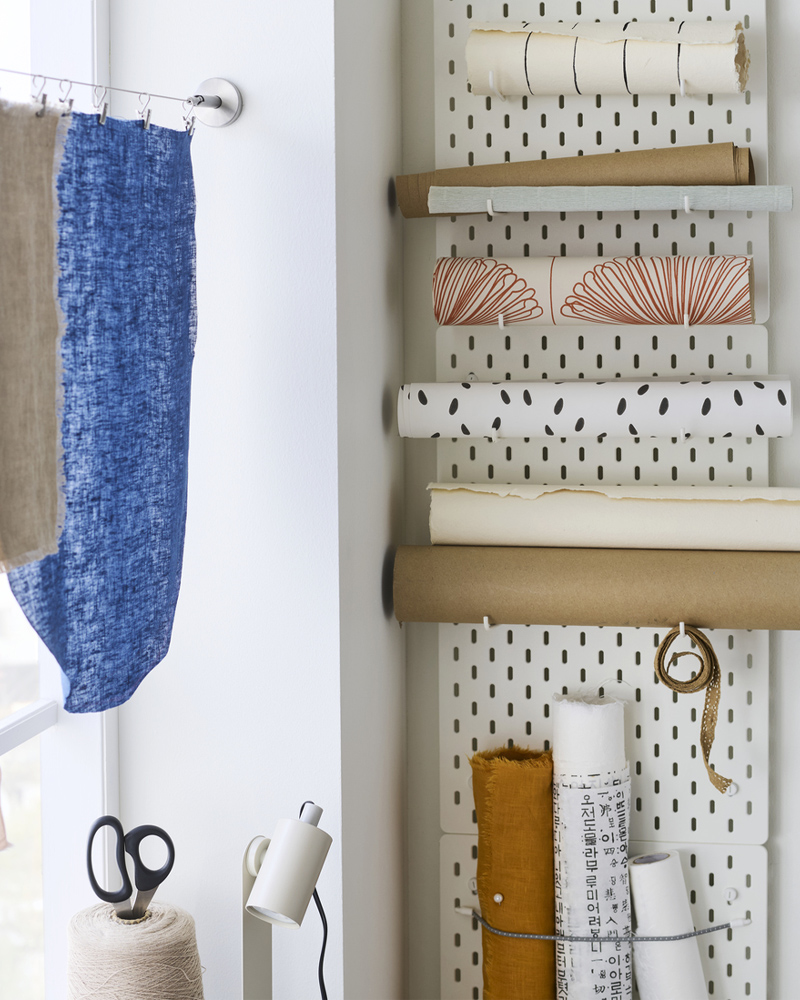 Three white SKÅDIS pegboards in a vertical row on a wall with hooks and elastic cord being used to hold rolls of paper.