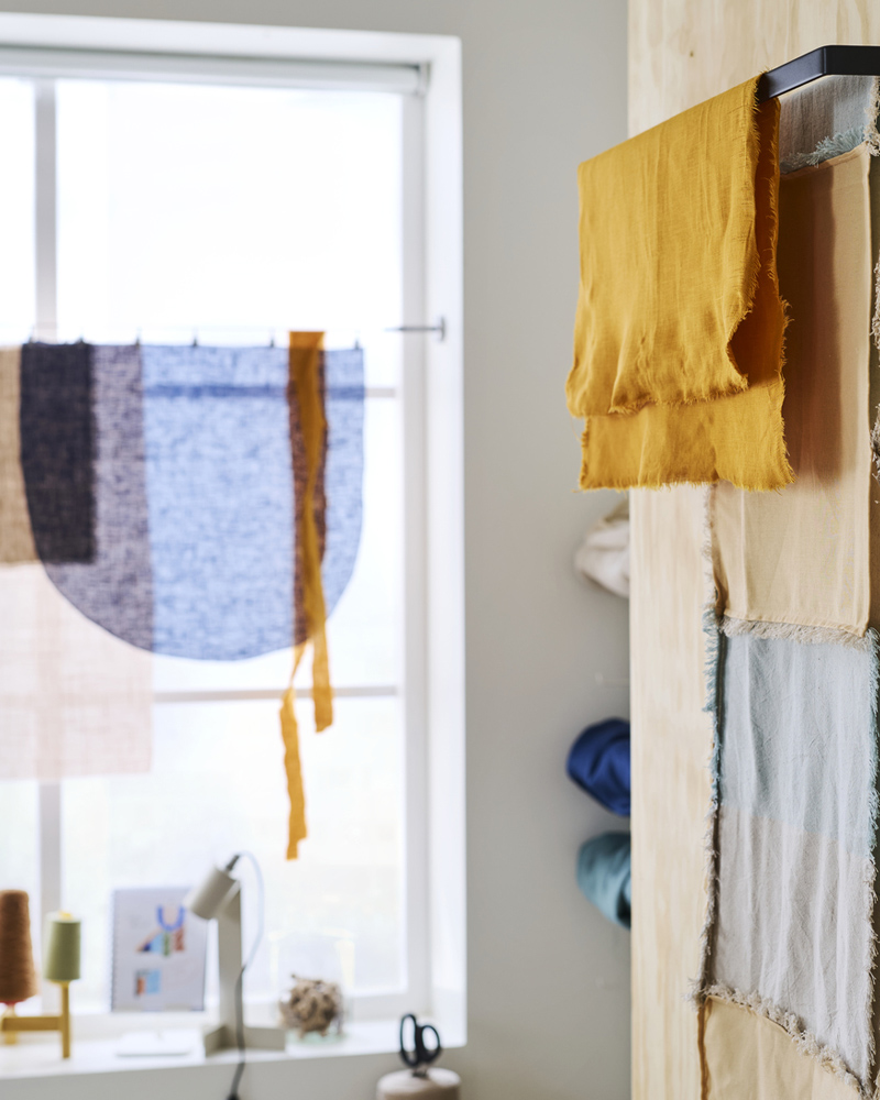 A black SKOGSVIKEN towel rail against a wooden wall holding different types of fabric near a desk with a FLOTTILJ lamp on it.