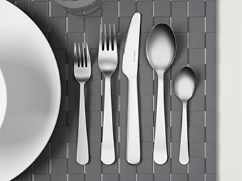Stainless steel cutlery set with the brand logo on the knife blade. It is placed on a gray woven placemat, next to a white plate.