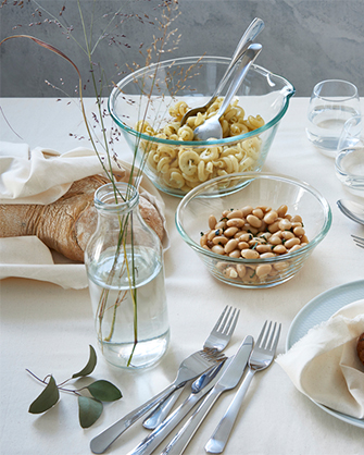 Dining table with a white tablecloth and pastel-colored IKEA plates, set with pasta salad and beans in glass bowls. Cutlery, bread, and a glass jar with branches are also on the table.