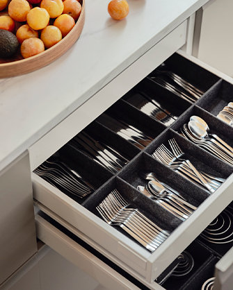 An open kitchen drawer with a gray felt organizer, showing multiple sets of stainless steel cutlery neatly arranged. To the left, a wooden bowl of fruit is visible on the countertop.