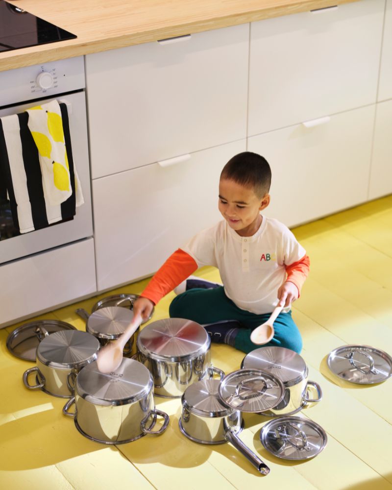 A child is sitting on a yellow kitchen fl oor, drumming with wooden spoons on IKEA 365+ cookware spread out on the floor.