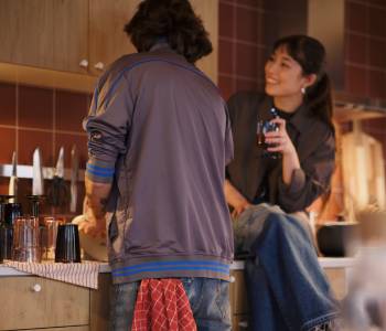 A person sits on a kitchen worktop, smiling at another person facing the worktop with a RINNIG tea towel in the back pocket.
