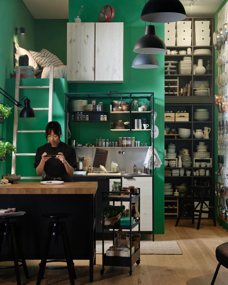 A person at a kitchen island photographs a plate of food. An ÄSPINGE kitchenette is placed by a green wall in the background.