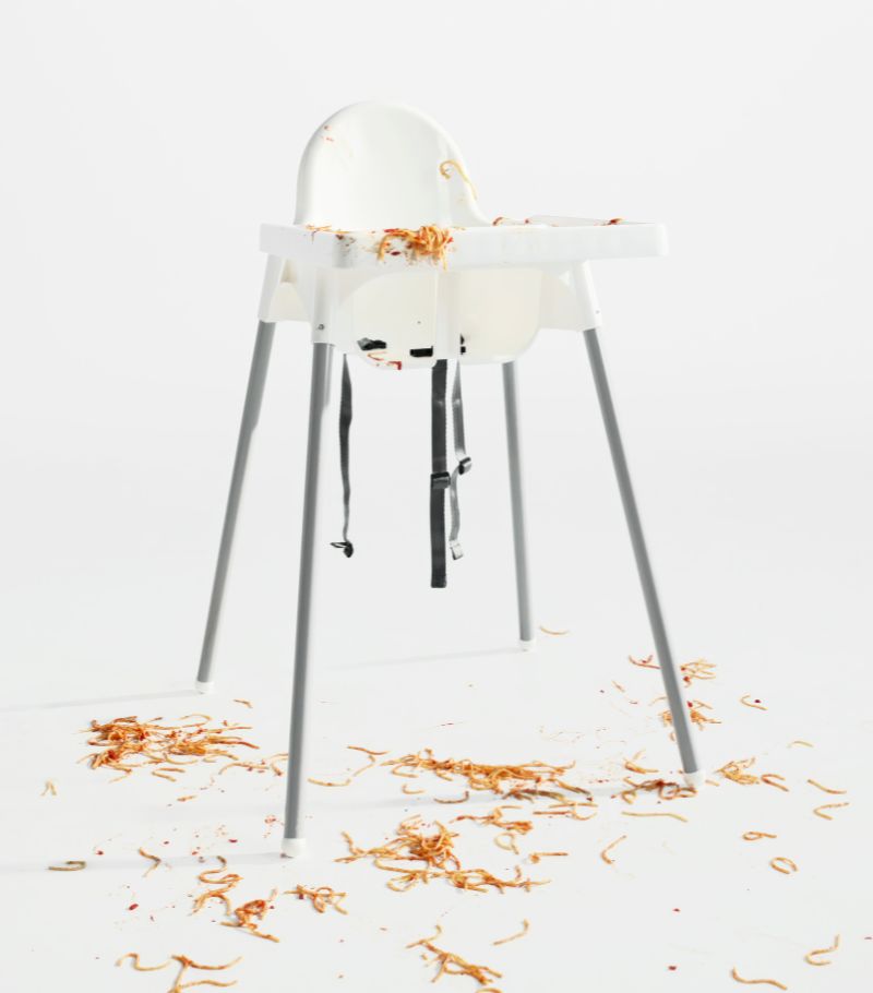An empty highchair with tray in a white space. The remains of a messy spaghetti meal cover the highchair and floor.