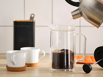 A close-up of a double-walled glass coffee press being filled with hot water. Two cups and a canister are next to it.