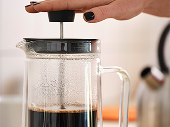 A close-up of a glass coffee press with the plunger being pushed down. A hand with black-painted nails is pressing the plunger.