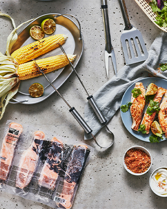 An assortment of barbecue food on a concrete countertop. It includes grilled corn, packaged fresh salmon, bowls with spices, and cooking utensils.