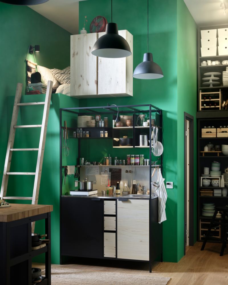 An ÄSPINGE kitchenette with utensils, spices, and more sits against a green wall next to a ladder leading to a sleeping loft.