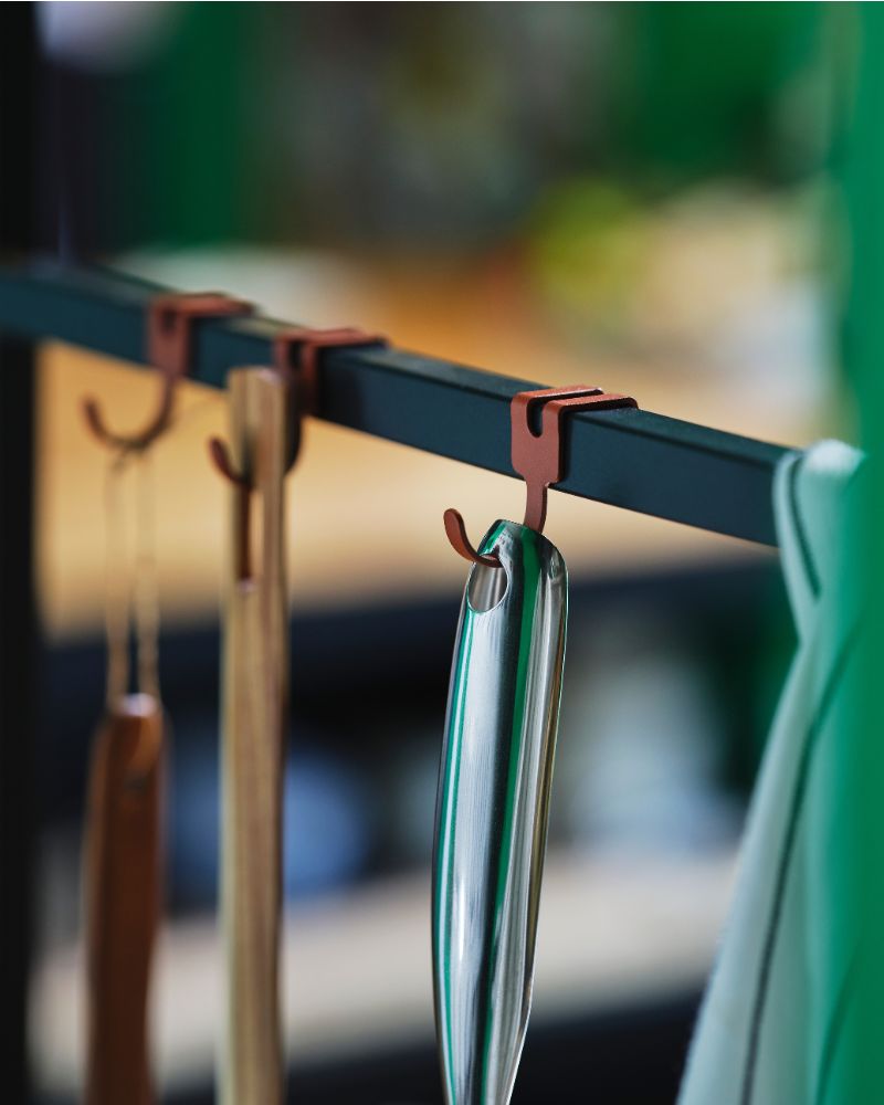 A close-up of various metal and wooden kitchen utensils hanging on reddish hooks in an ÄSPINGE kitchenette.