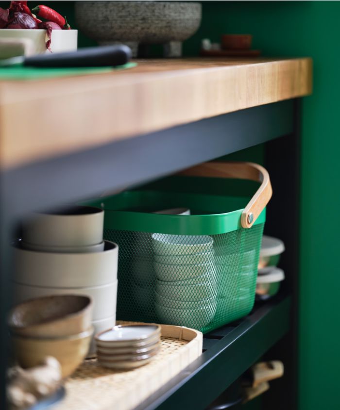 A dark green RISATORP basket with a wooden handle sits on a shelf of a kitchen island next to a tray, plates, and bowls.