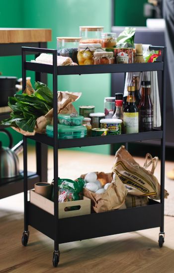 The shelves of a black NISSAFORS trolley are teeming with fresh vegetables, eggs, jars and cans of food, and various bottles.