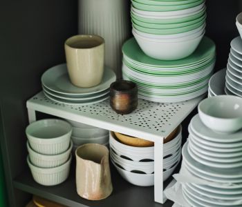 A white VARIERA shelf insert sits in a shelving unit. There are stacks of bowls and plates on, under, and next to it.