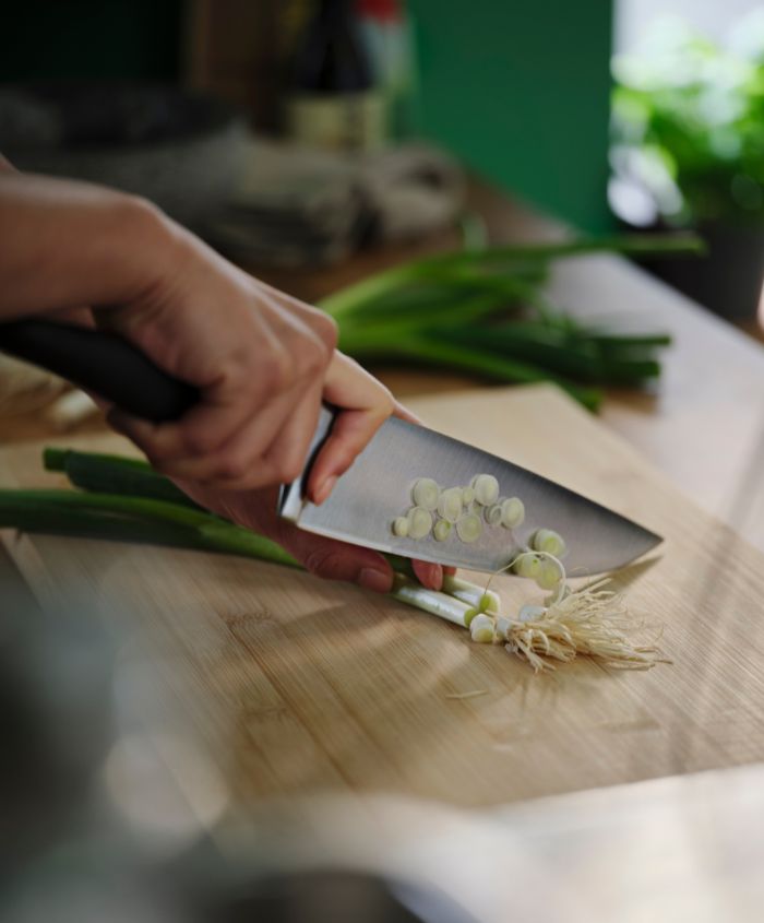 Two hands are holding and cutting vegetables on a bamboo APTITLIG chopping board with a black VÖRDA cook’s knife.