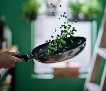 A hand is holding a stainless steel MIDDAGSMAT frying pan with a non-stick coating, tossing the veggies in it up in the air.
