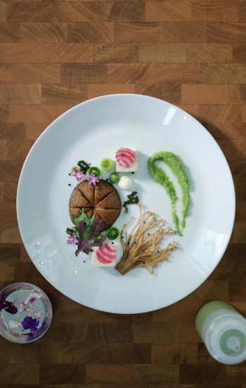 A white OFTAST plate is seen from above on a wooden surface. Food is beautifully arranged on it in a fine dining style.