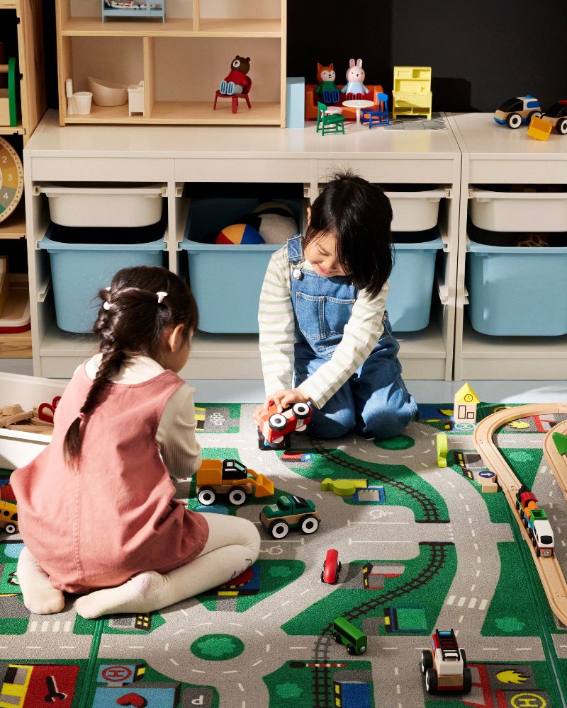Two kids play on a colorful car rug with toys. The room has light wood furniture and blue and white storage bins.
