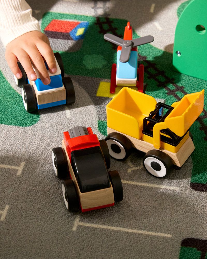 A child's hand plays with toy cars: a red and black truck, a yellow excavator, and a blue helicopter.
