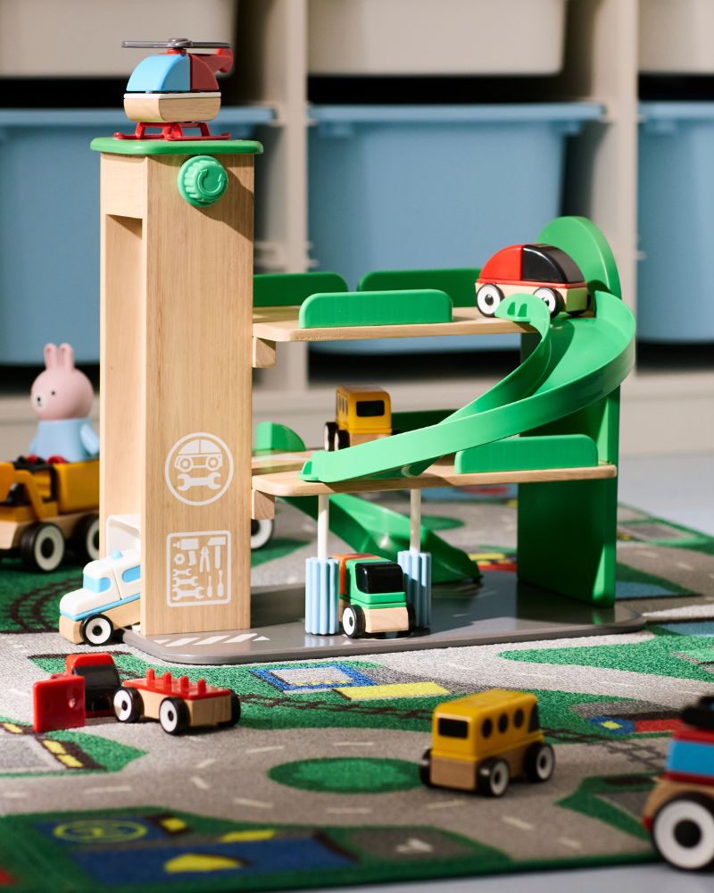 A multi-level wooden toy parking garage with a green ramp, a red car, a helicopter, and other toy vehicles.