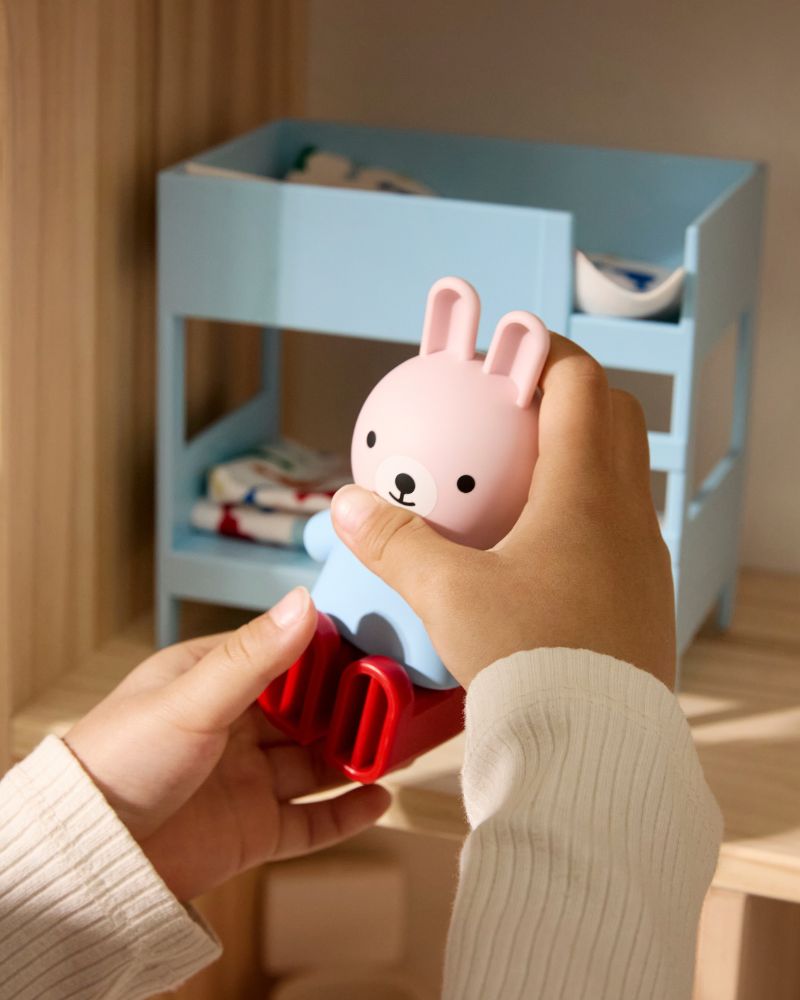 A child's hands hold a pink and blue rabbit toy. In the background, there's a light blue dollhouse bunk bed.