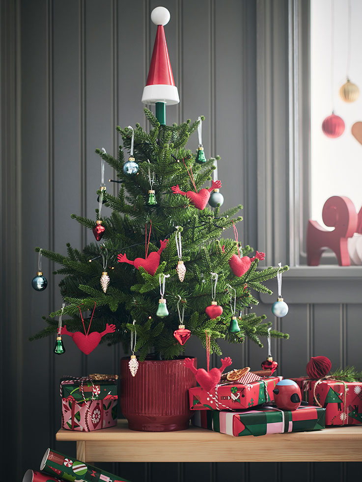 A small potted Christmas tree is decorated with playful VINTERFINT tree ornaments and placed on a bench with presents on it.