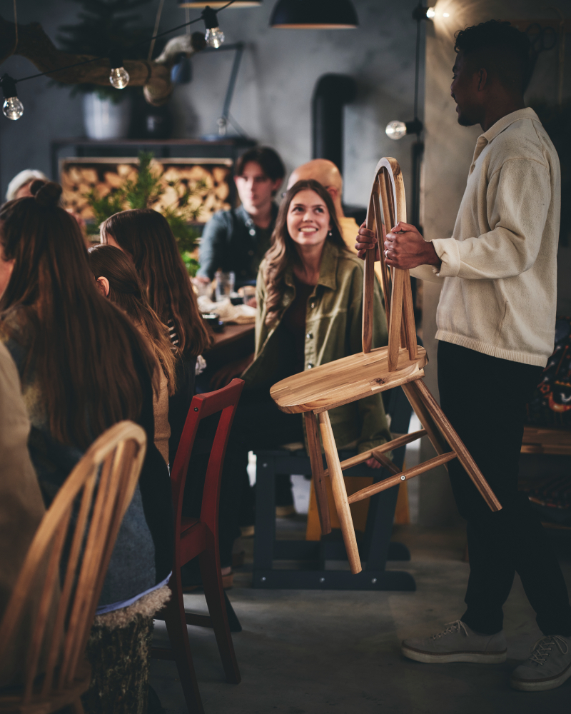 A young man brings a wooden dining chair to a long table surrounded by seated dinner guests.