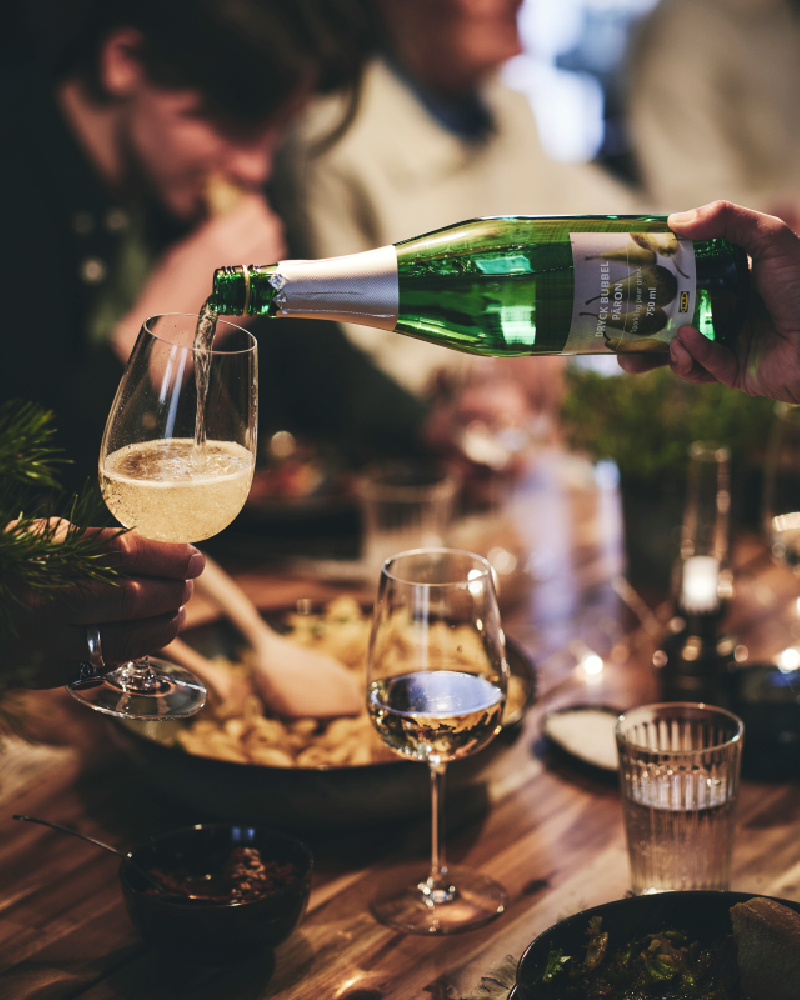 A guest at a dinner party pours a glass of sparkling drink into another guest’s wine glass.