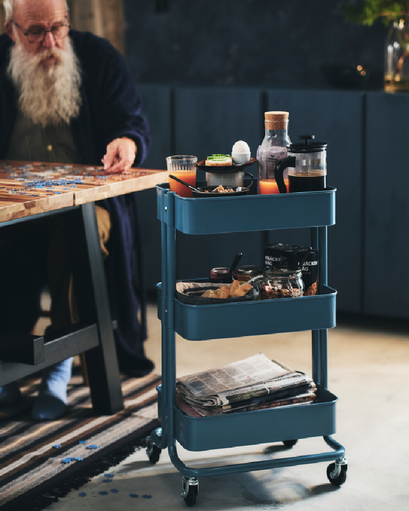 An elderly man with a long beard lays a jigsaw puzzle on his dining table. Beside him is a RÅSKOG trolley with his breakfast.