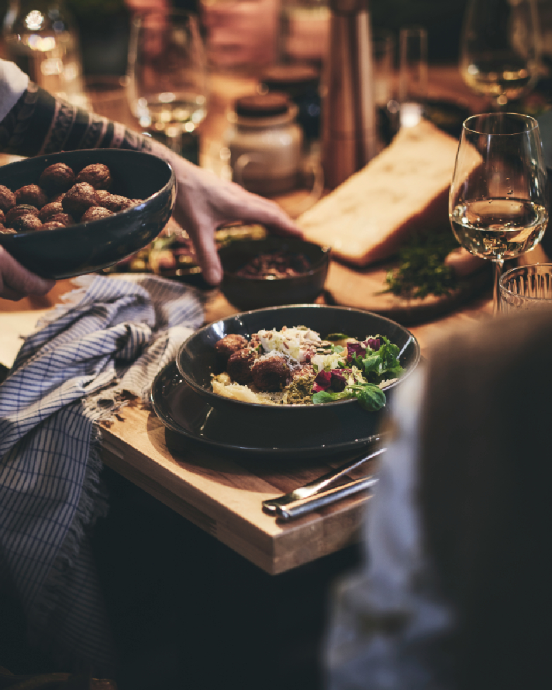 A dinner guest at a long table passes a bowl of HUVUDROLL plant balls to the guest sitting beside them.