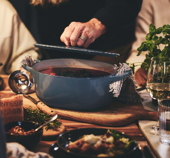 An elderly man with a long beard lifts the lid on a casserole dish while dinner guests seated at the table look on.
