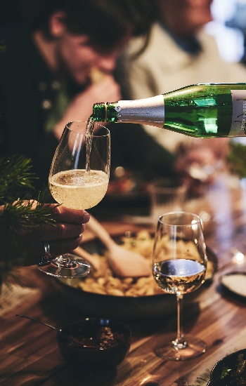 A guest at a dinner party pours a glass of sparkling drink into another guest’s wine glass.