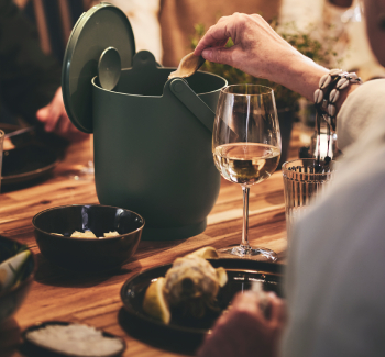 An elderly woman sitting at a dinner party places her artichoke waste in a stylish on-table waste bin