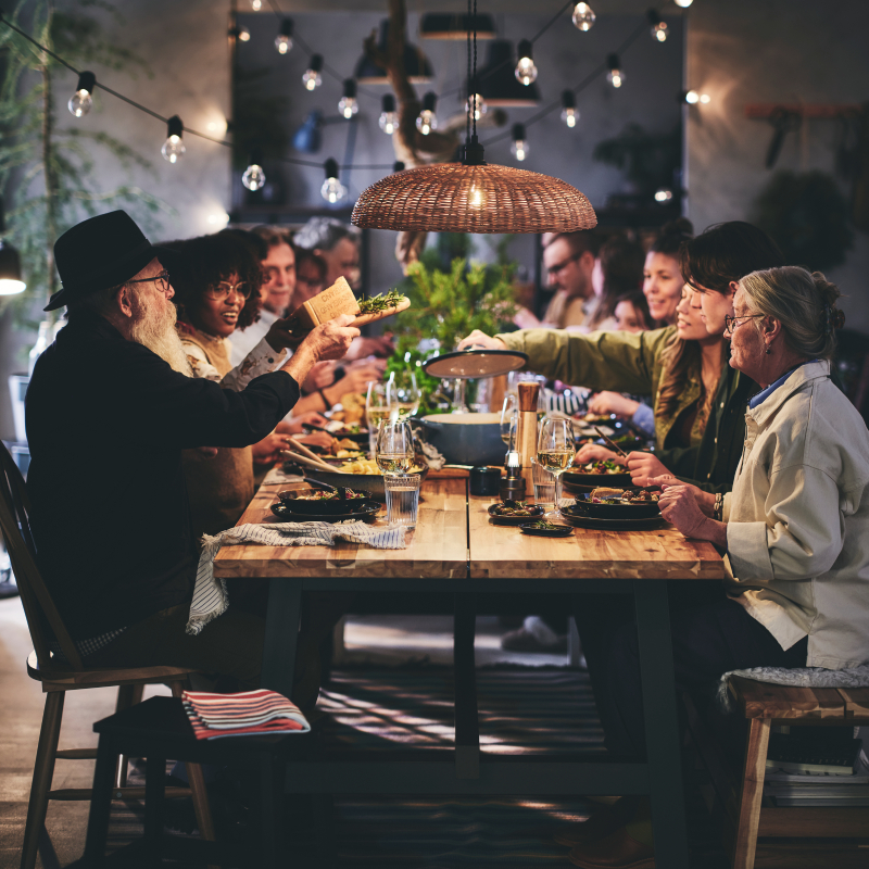 An elderly couple seated at the end of a long table surrounded by seated dinner guests of all ages.
