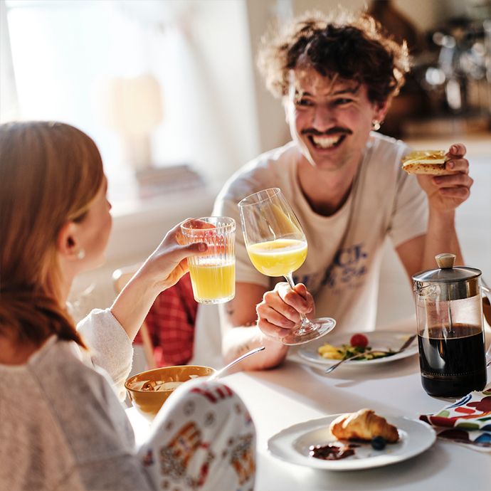 Couple smiling and toasting with orange juice at a round white dining table, set with plates, a croissant, and a French press coffee maker.