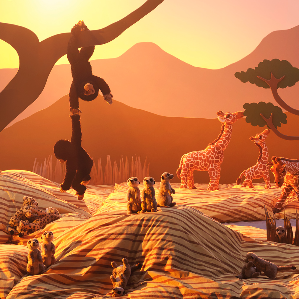 A sunlit savannah made from cardboard cutouts and duvet covers and pillowcases is filled with SANDLÖPARE soft toy animals.
