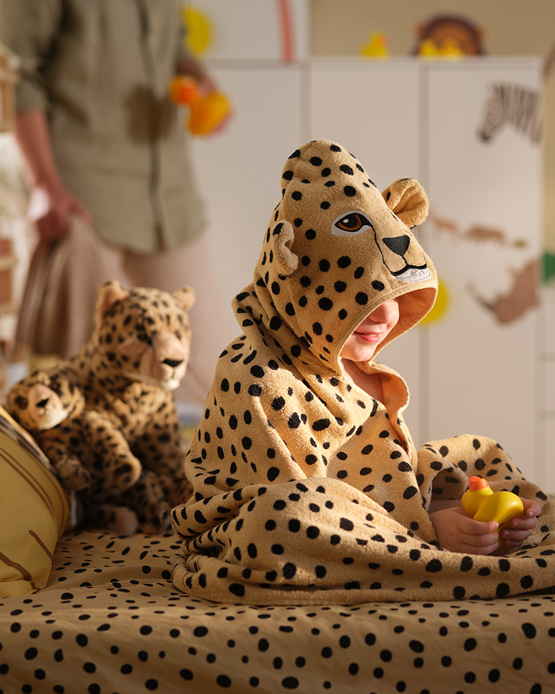 A child in a SANDLÖPARE towel with hood with cheetah pattern sits on a bed surrounded by soft toy cheetahs and a rubber duck.
