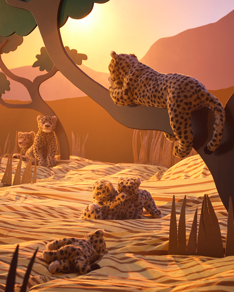 A group of beige SANDLÖPARE soft toy cheetahs and cheetah cubs are playing and sleeping in a sunlit savannah landscape.