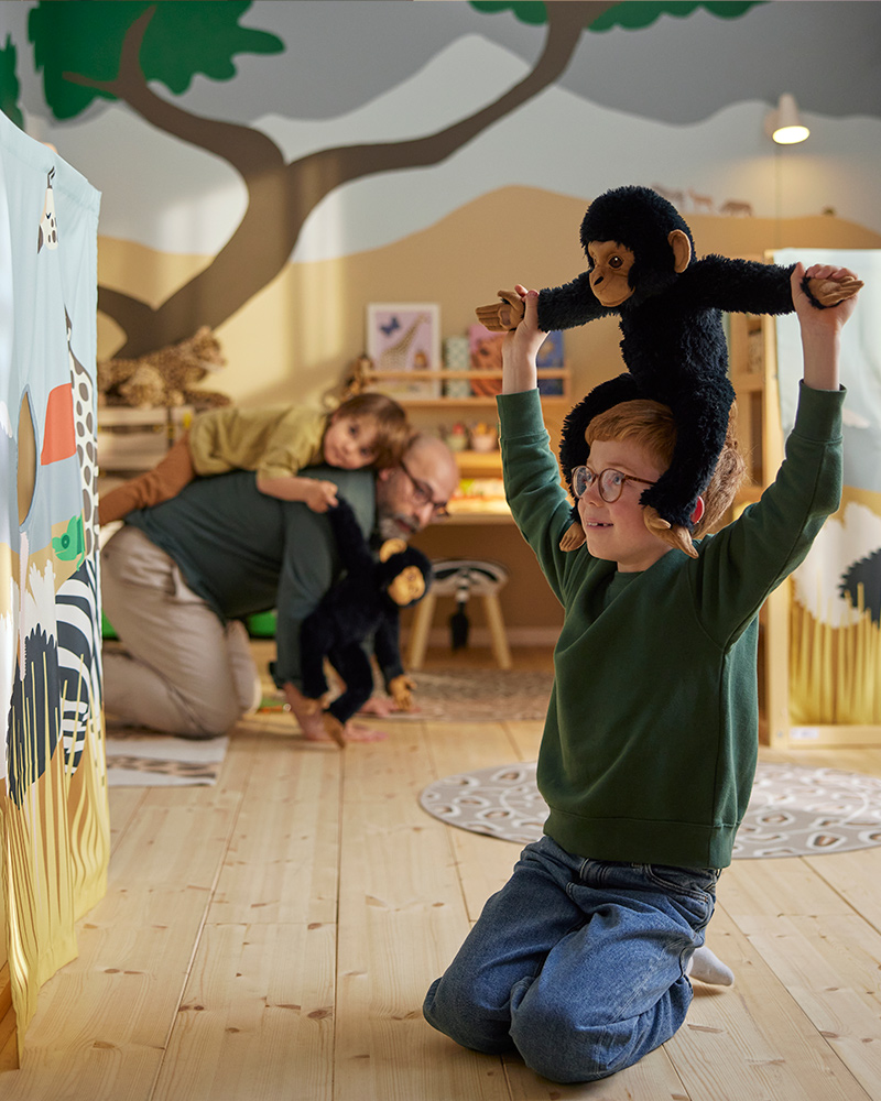 A child sits on their knees in a savannah themed room while carrying a SANDLÖPARE soft toy chimpanzee on their shoulders.