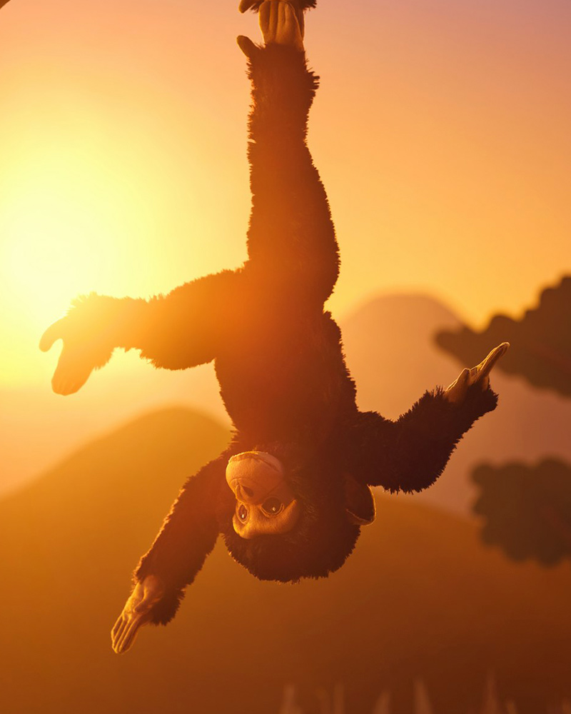 A black SANDLÖPARE soft toy chimpanzee hangs by its feet in a sunny savannah landscape.