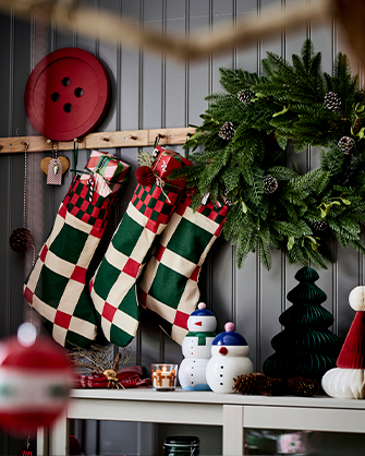 Pared de listones grises decorada con tres calcetines navideños de tela a cuadros rojo, verde y beige, colgados de un estante sobre figuras de decoración de papel y cerámica.